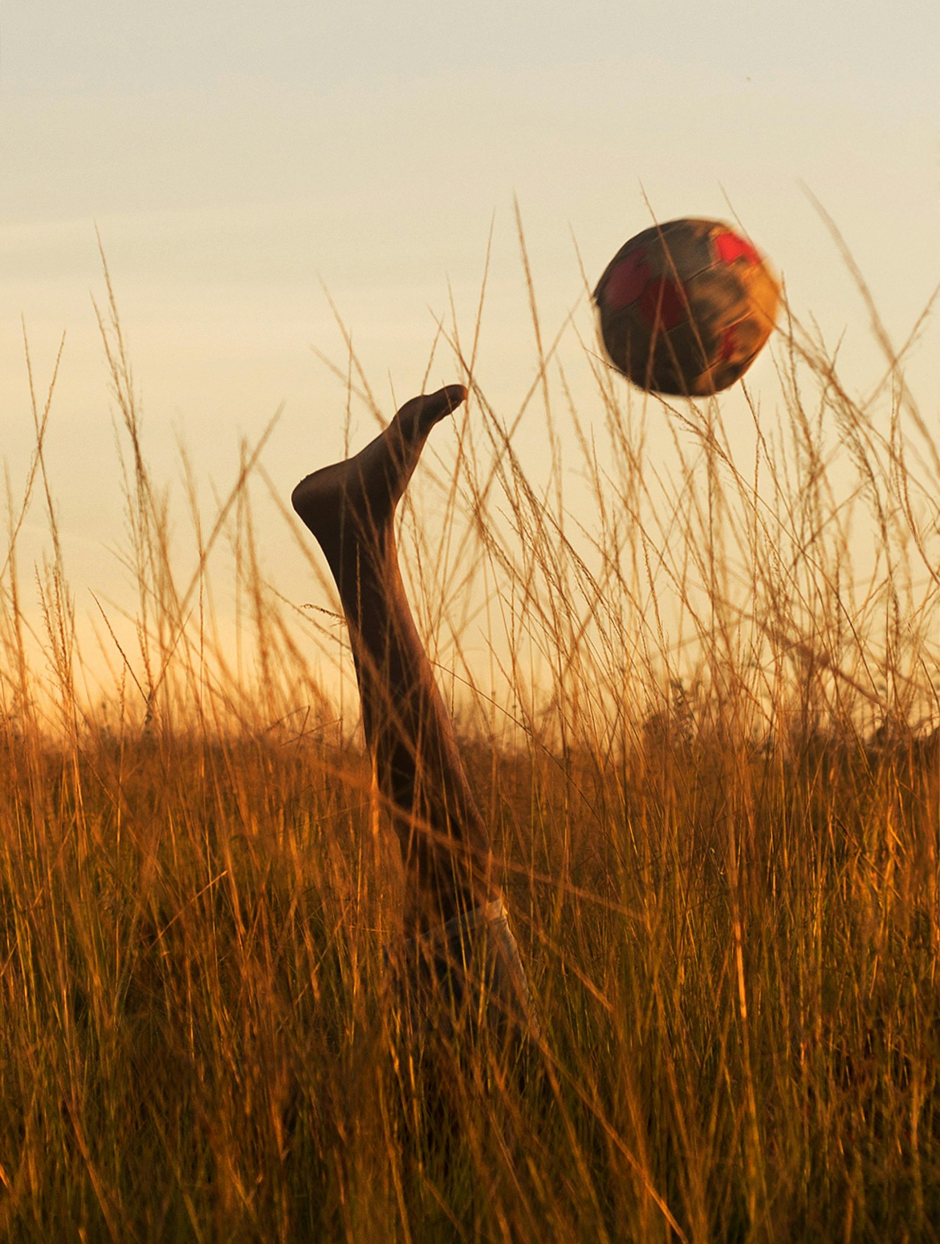 hero-img-01 A young boy plays soccer in tall grass at sunset in Rio de Janeiro, embracing freedom and joy.