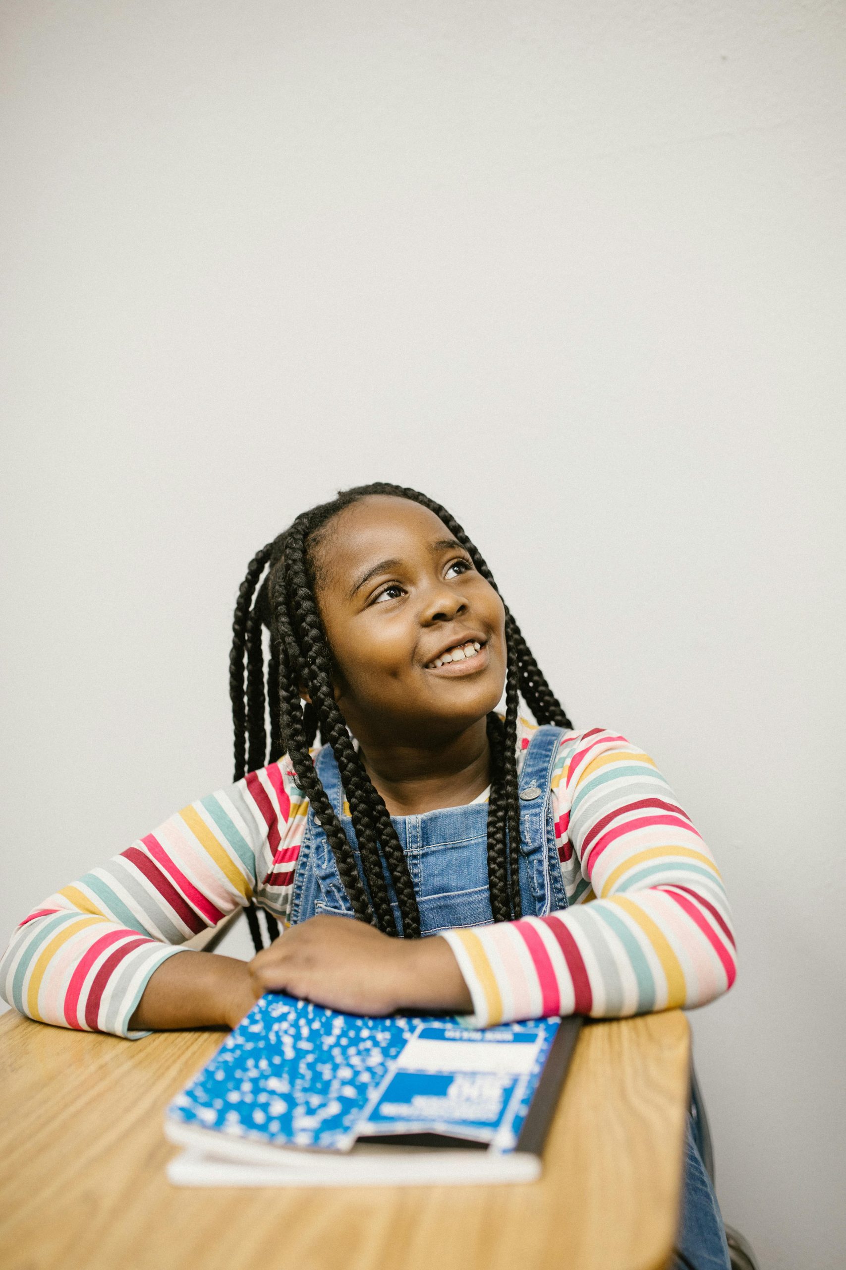 our-services-1 A cheerful young black girl smiling while sitting at a desk with a notebook in classroom.